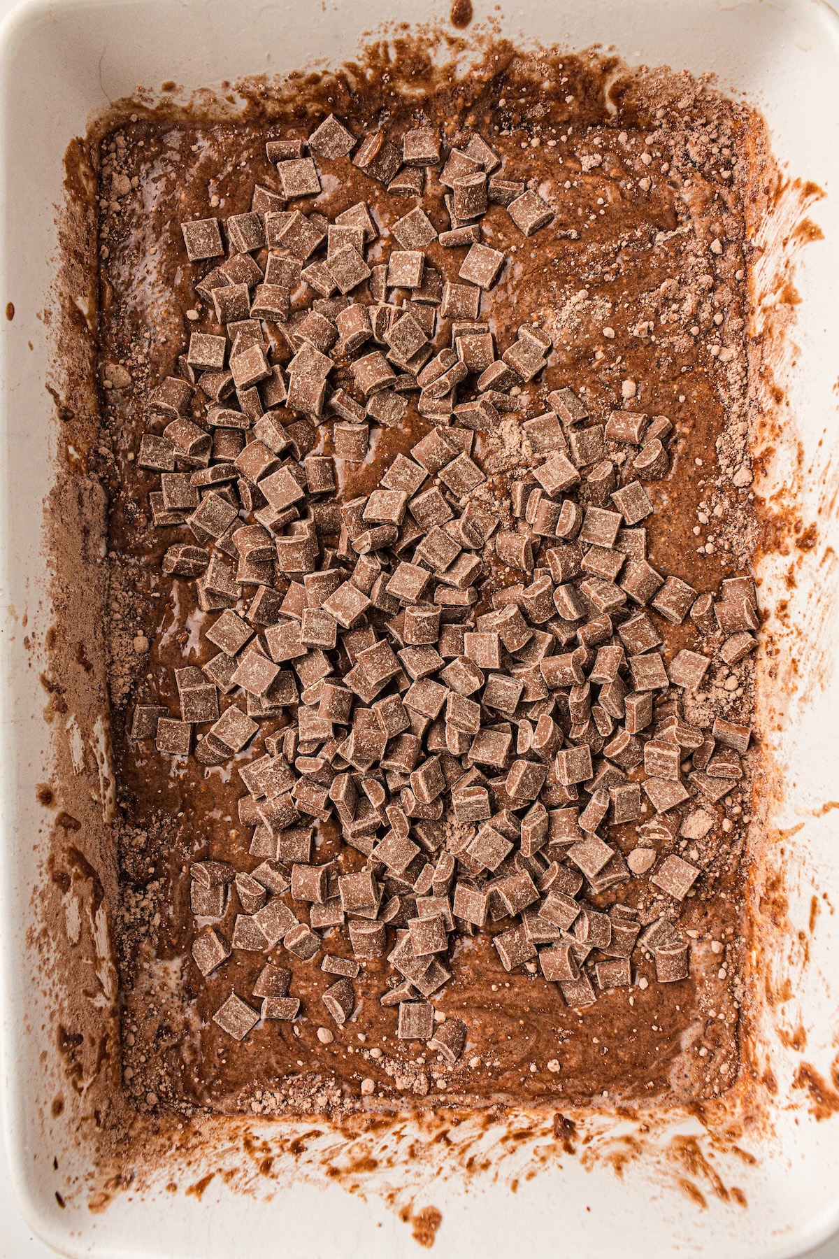 Top-down view of a white rectangular baking dish filled with a thick chocolate mixture topped with scattered milk chocolate chunks. The mixture appears wet and textured, with traces of cocoa powder visible, suggesting the dessert is ready for baking.