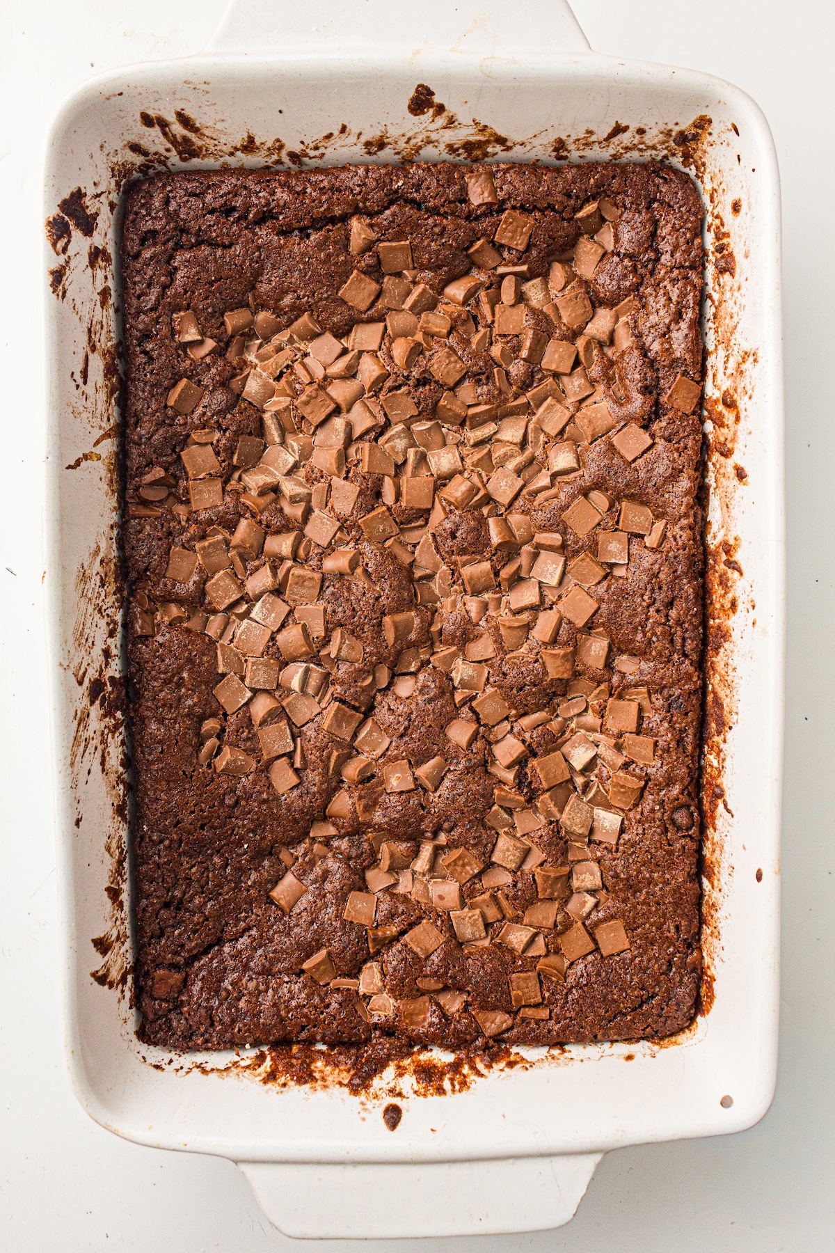 Overhead view of a freshly baked chocolate brownie in a white rectangular baking dish. The brownie has a crinkly top and is generously topped with melted milk chocolate chunks, creating a glossy, indulgent appearance.