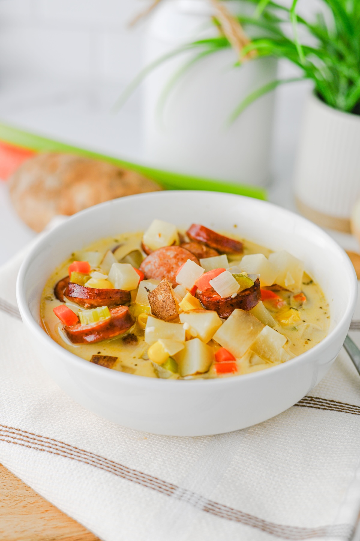 Front view of a full bowl of kielbasa potato soup with fresh vegetables and a potato blurred in the background.