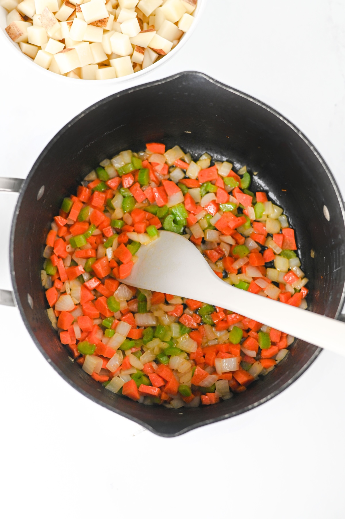 Diced onion, carrot, and celery saut&eacute;ing in a pot with a wooden spoon, potatoes waiting nearby.
