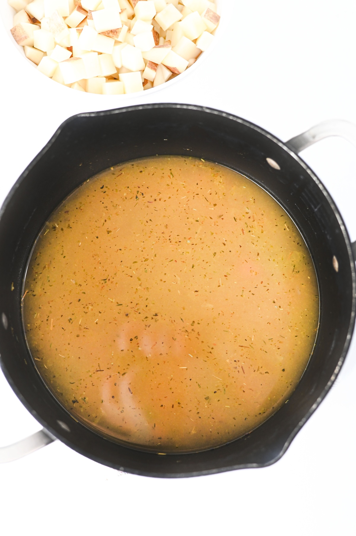 Chicken broth and seasonings simmering in a pot, with diced potatoes ready to be added.