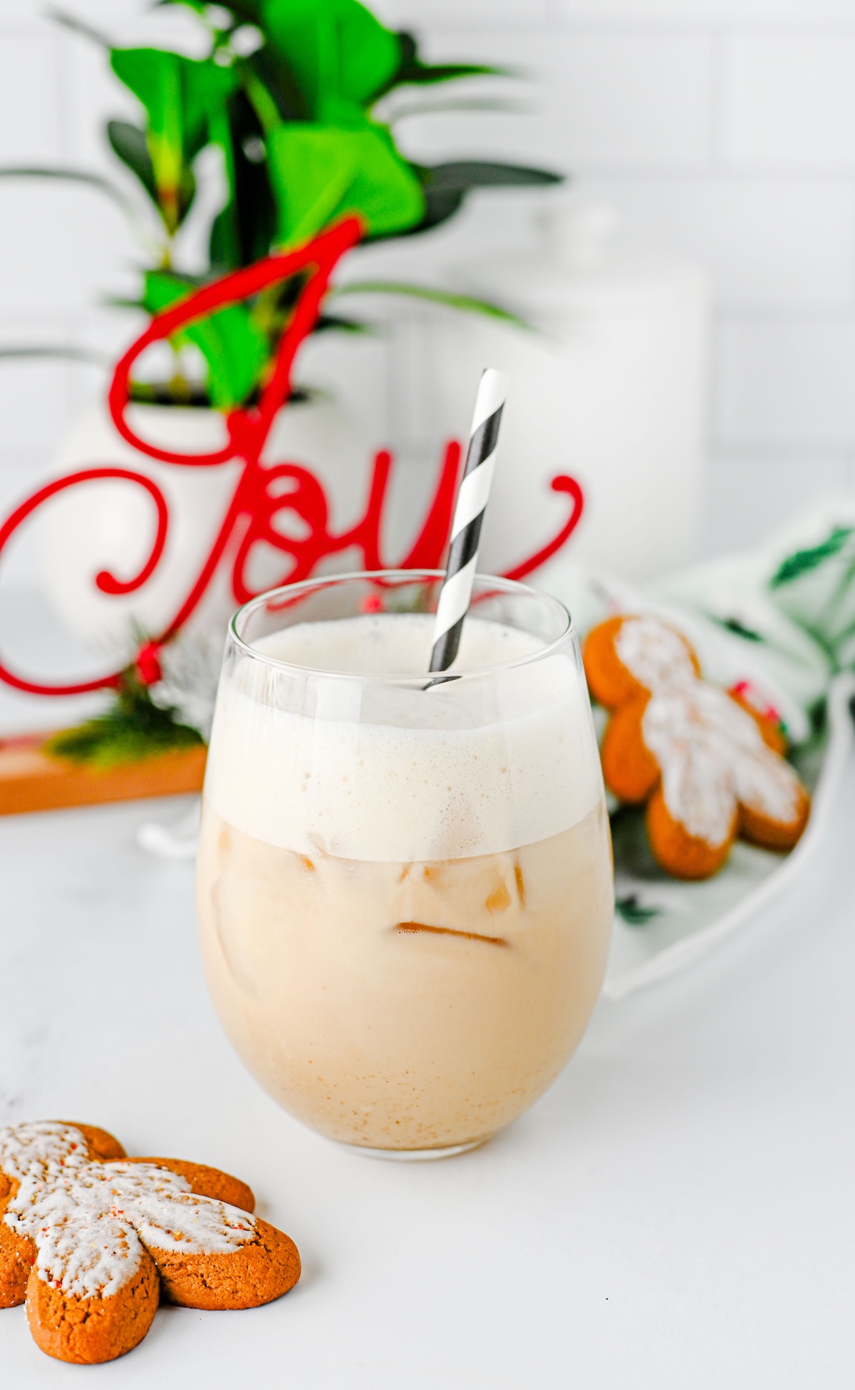 Iced gingerbread chai latte with frothy oat milk foam and a striped straw next to gingerbread cookies and a red Joy sign