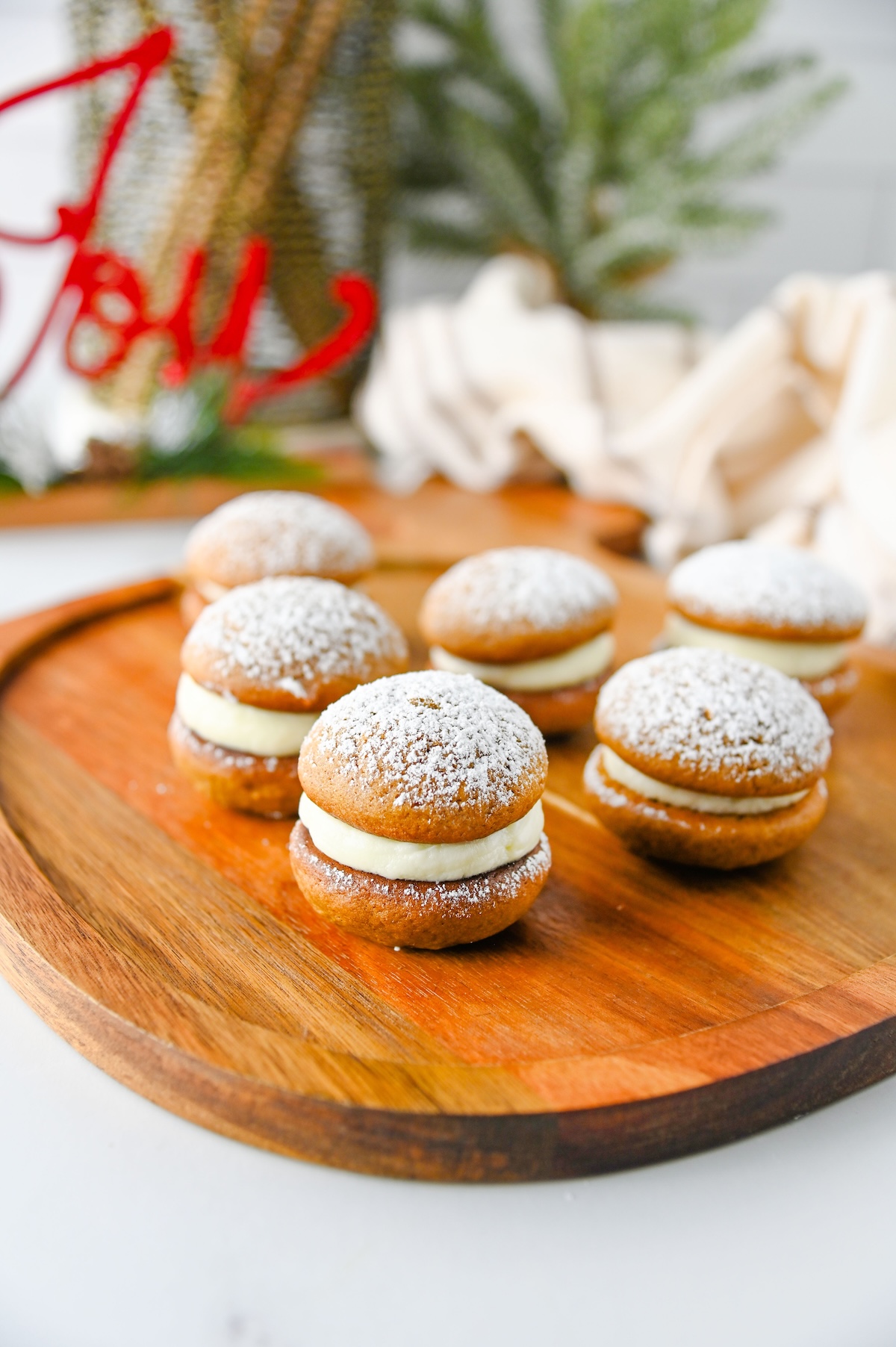 Gingerbread whoopie pies with cream cheese filling and powdered sugar on a wooden board with holiday decor behind them