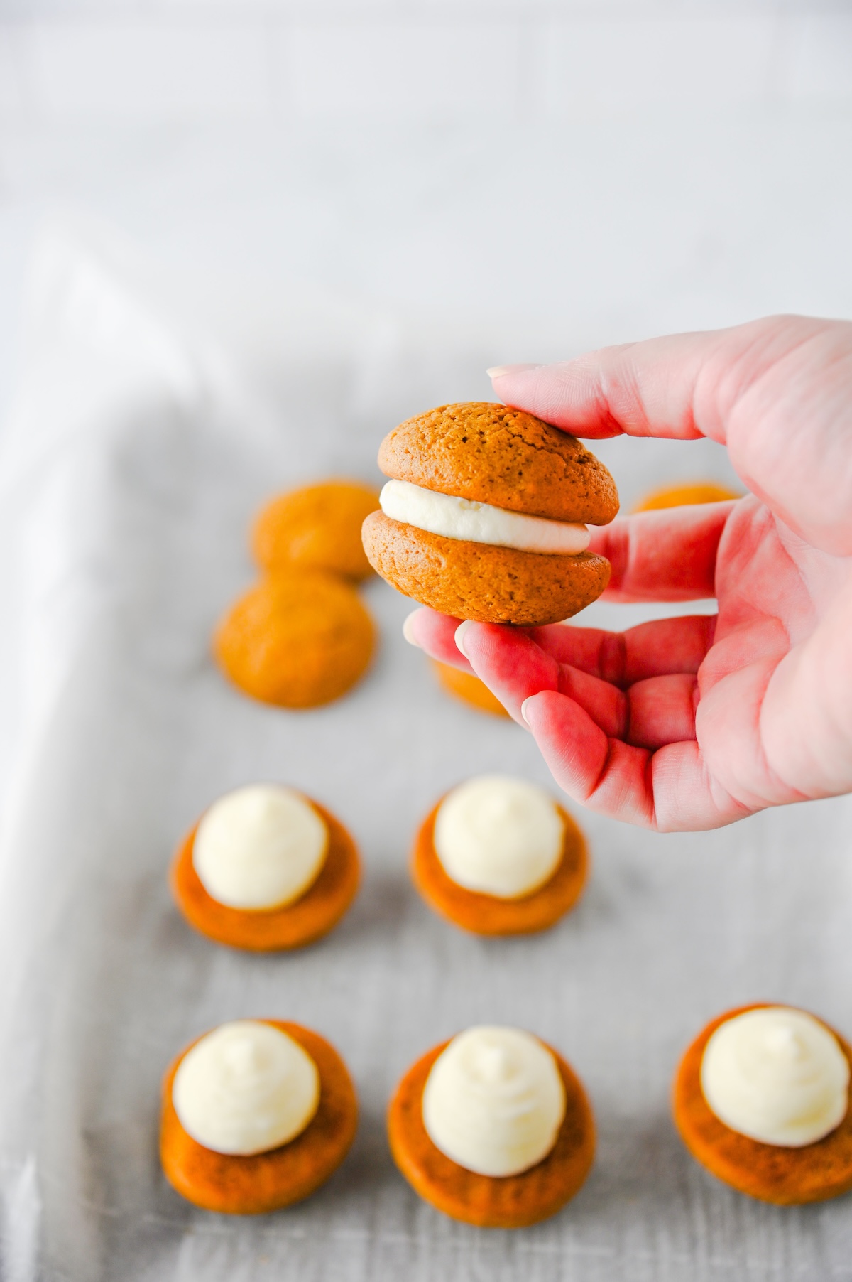 Hand holding an assembled gingerbread whoopie pie over a baking sheet of cookies topped with filling