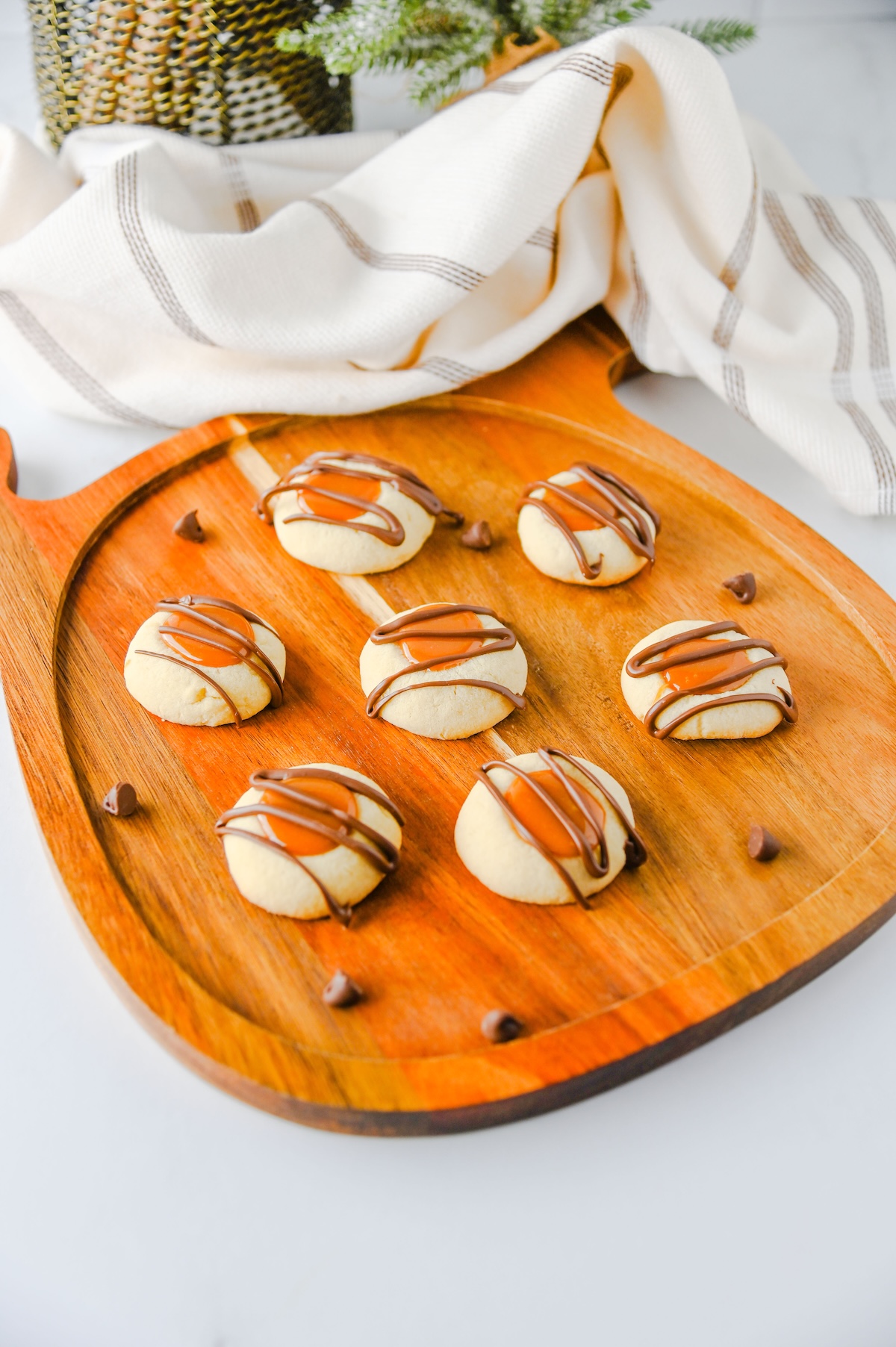 Twix thumbprint cookies on a pumpkin-shaped wooden board with a striped kitchen towel and greenery