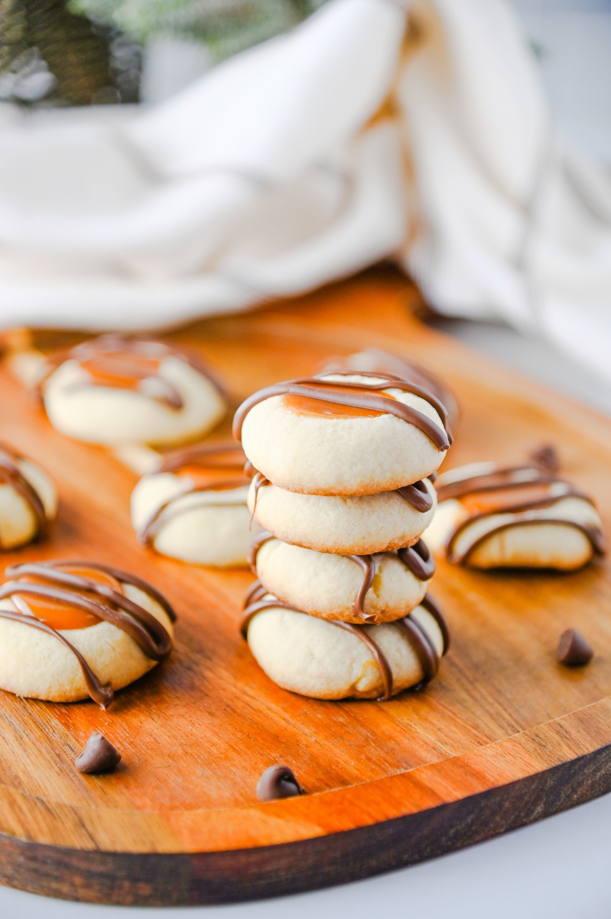 Stack of Twix thumbprint cookies on a wooden board surrounded by more cookies and chocolate chips