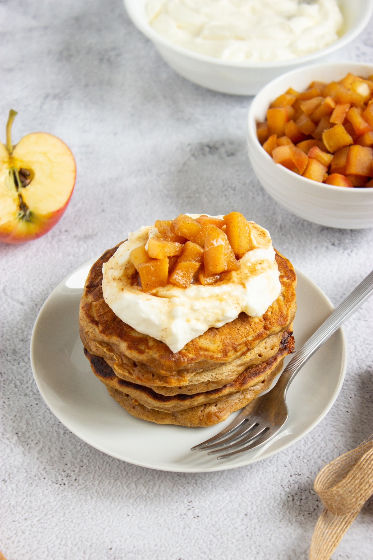 Stack of apple cinnamon pancakes topped with whipped cream and caramel apple compote on a white plate with a fork and a halved apple