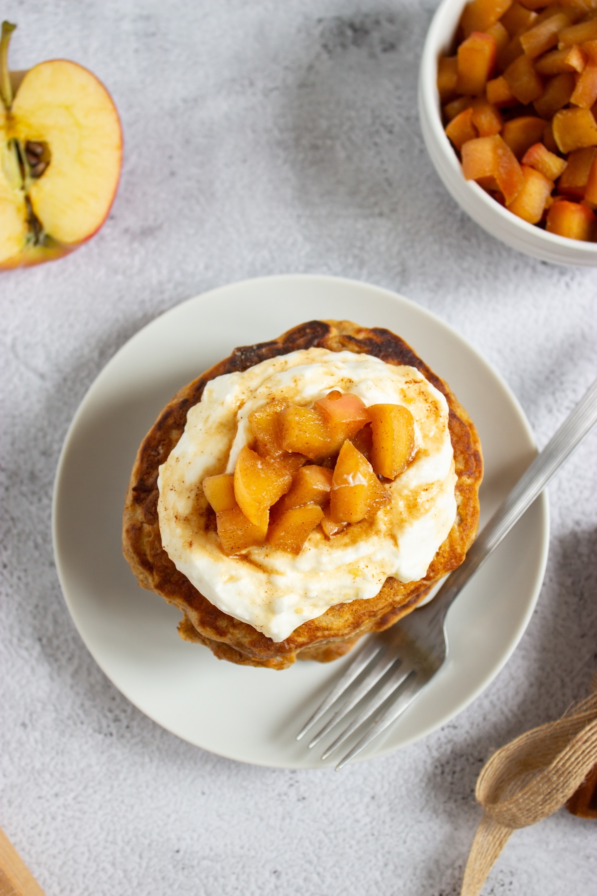 Overhead view of apple cinnamon pancakes topped with whipped cream and caramel apple compote on a white plate beside a bowl of compote and a halved apple