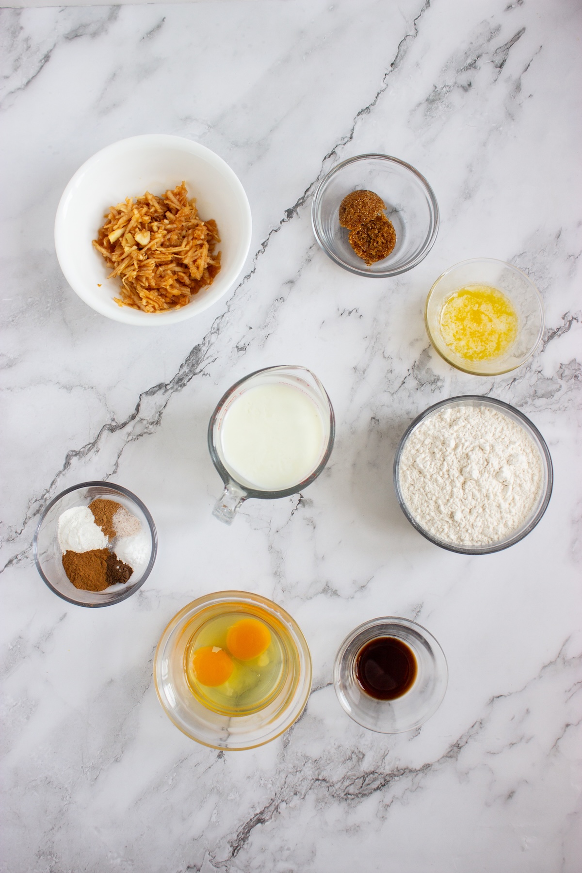Ingredients for apple cinnamon pancakes in bowls on a marble counter, including flour, buttermilk, eggs, grated apple, melted butter, vanilla, and spices