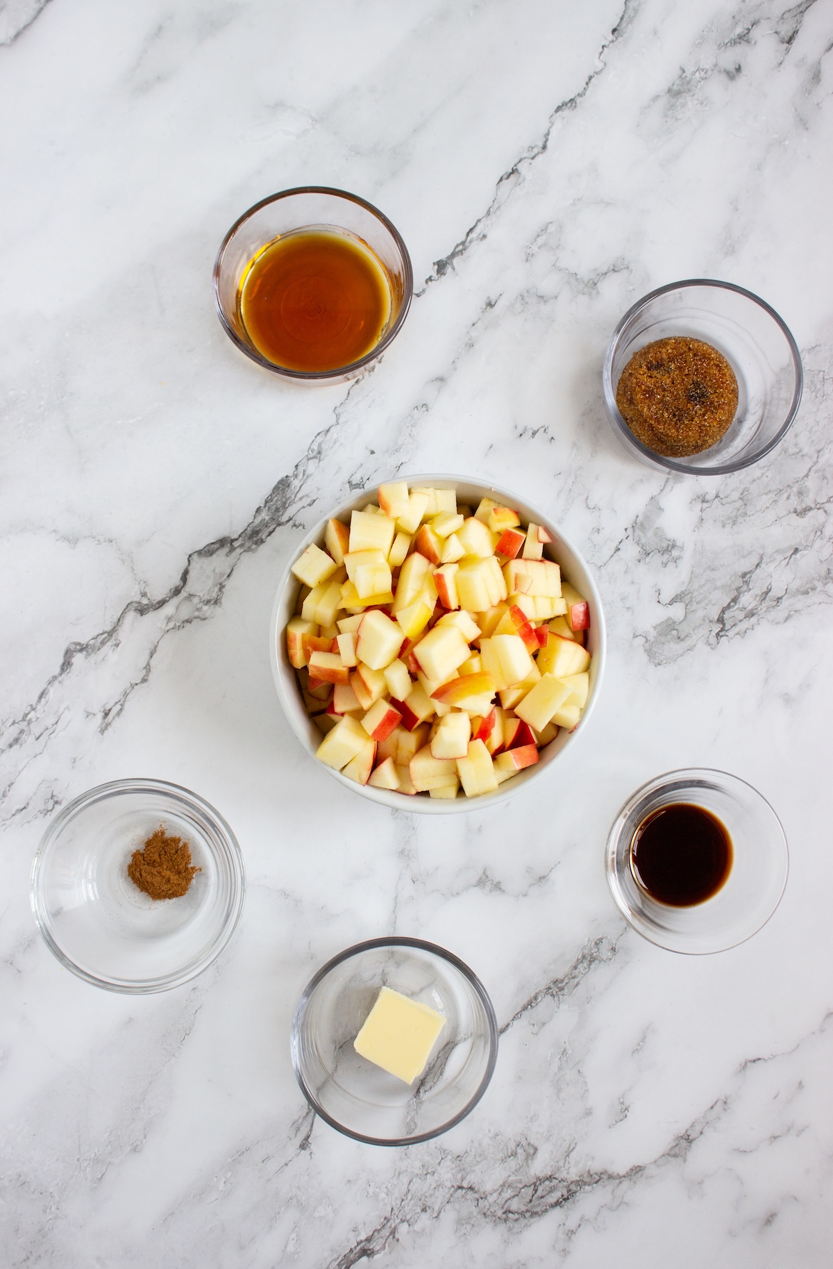 Ingredients for caramel apple compote on a marble counter, including diced apples, butter, brown sugar, maple syrup, cinnamon, and vanilla
