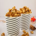Brown butter maple popcorn in two striped paper cups with cinnamon sticks and fall leaves
