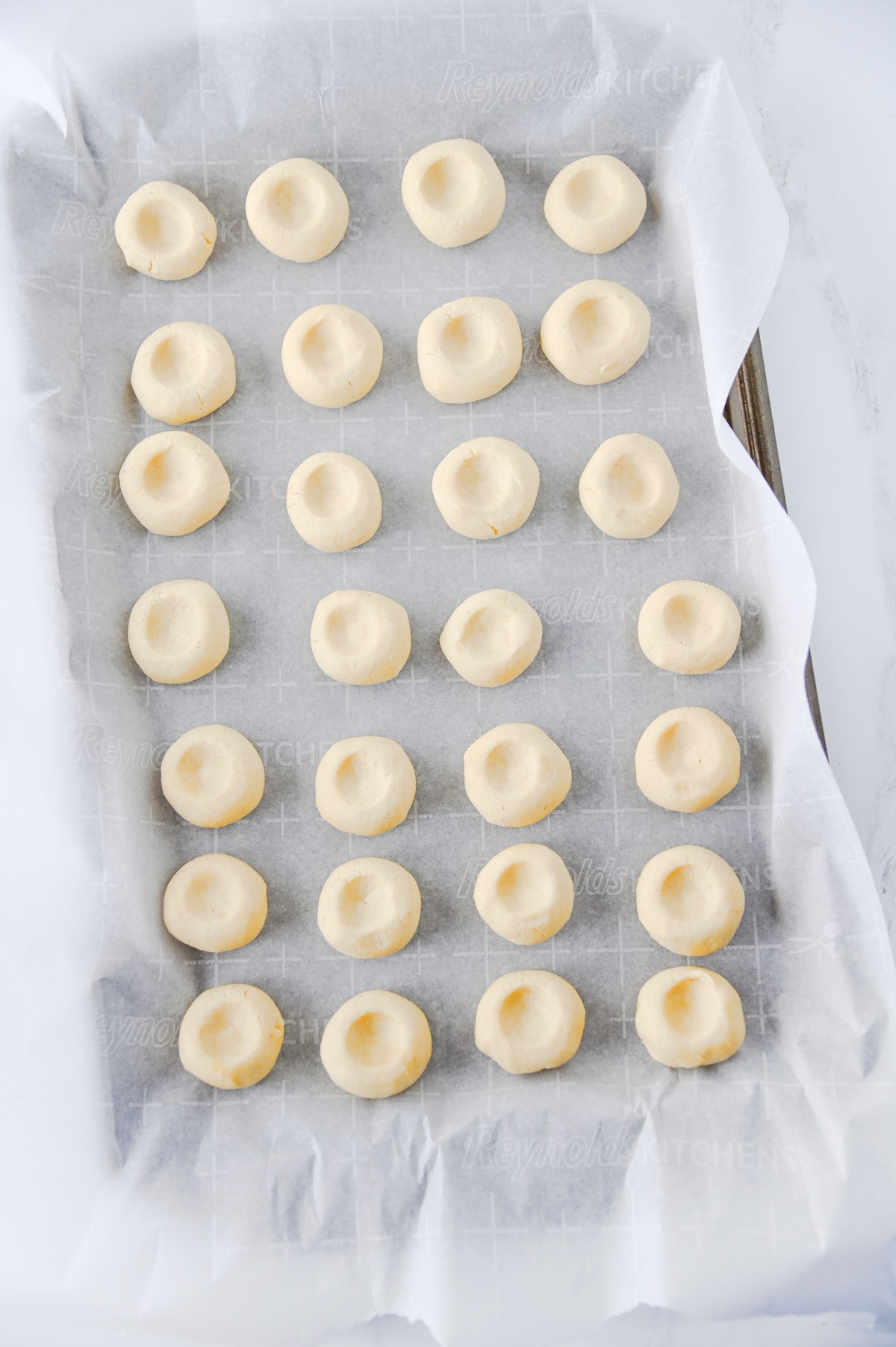 Cookie dough balls pressed with thumbprint indents on a parchment-lined baking sheet