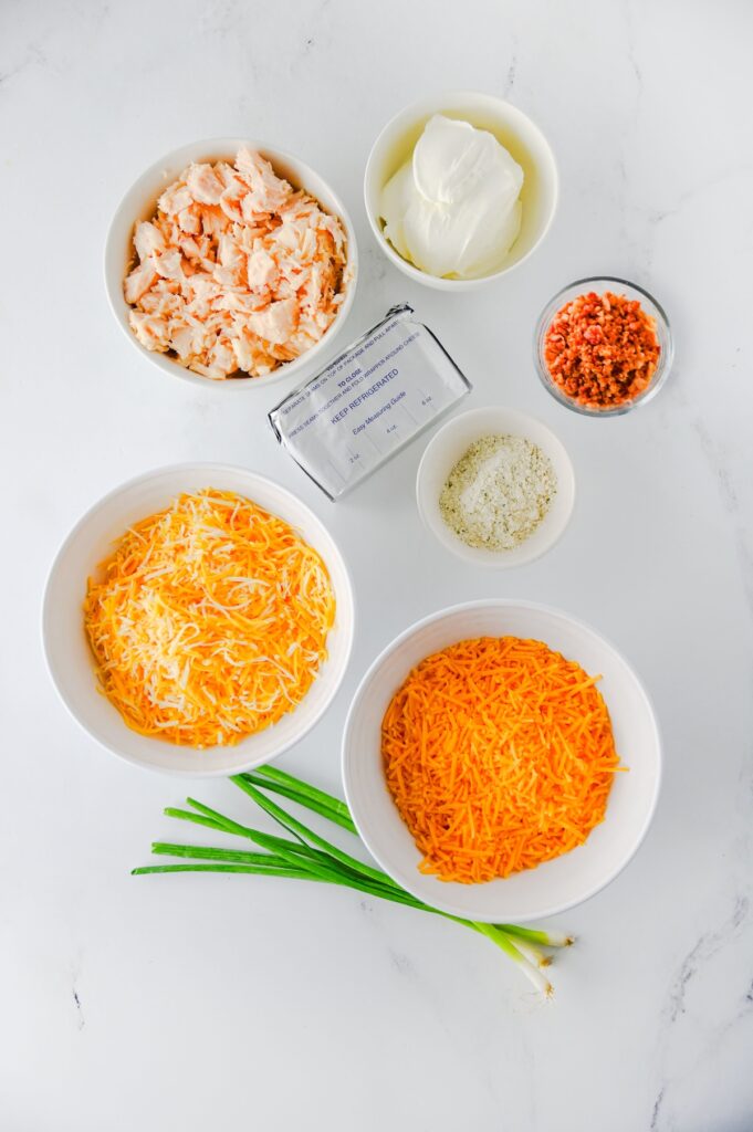 Crack chicken dip ingredients in separate bowls: canned chicken, sour cream, bacon pieces, ranch seasoning, Colby jack, sharp cheddar, and green onions